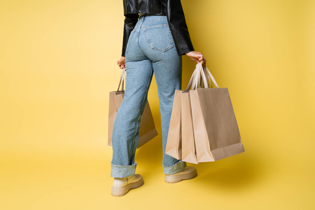黄色のショッピングバッグを持つスタイリッシュなレザージャケットとデニムジーンズでアフリカ系アメリカ人女性のクロップドビュー - 写真・画像