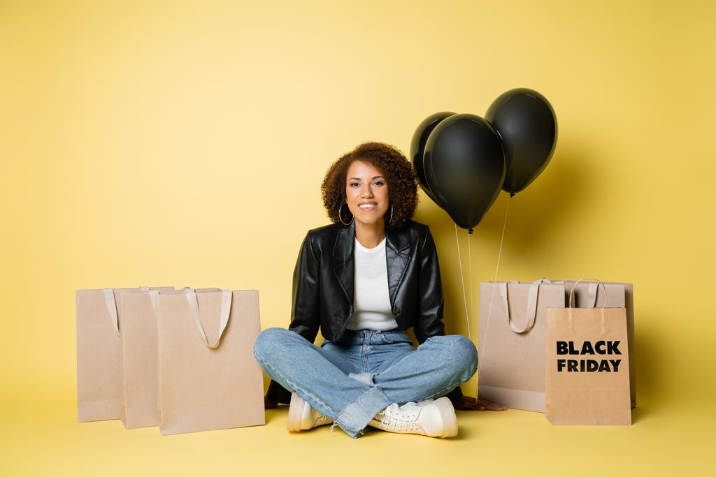 pleine longueur de souriante femme afro-américaine en veste en cuir assis près de sacs à provisions noir vendredi et ballons sur jaune - Photo, image
