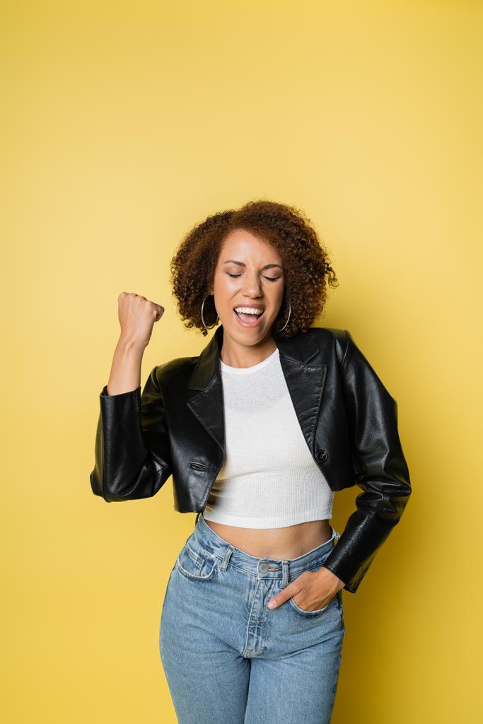 excited african american woman in stylish leather jacket and jeans posing on yellow - Photo, Image