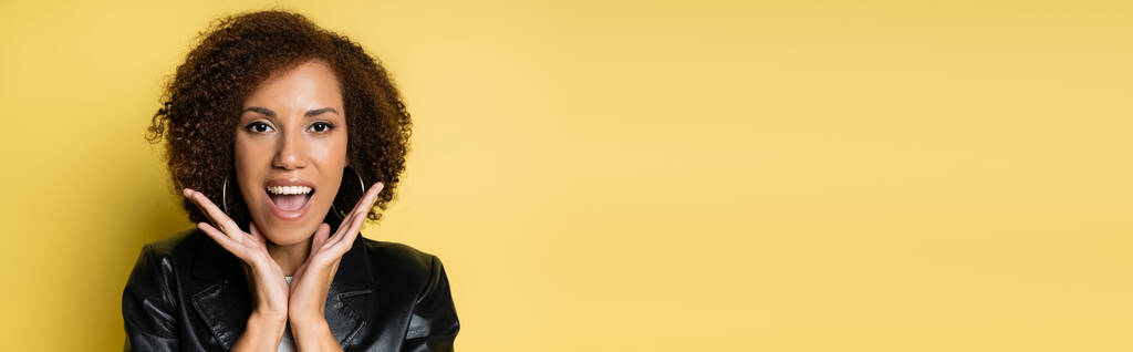 zdumiona Afroamerykanka w stylowej skórzanej kurtce gestykulując izolowane na żółtym, sztandarze - Zdjęcie, obraz