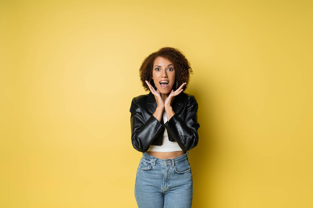amazed african american woman in stylish leather jacket gesturing while standing on yellow - Photo, Image