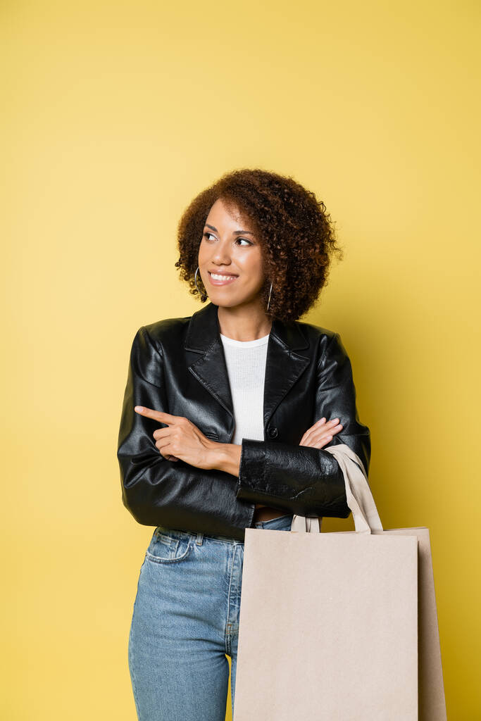 happy african american woman in stylish leather jacket holding shopping bags with purchases and pointing with finger on yellow - Photo, Image