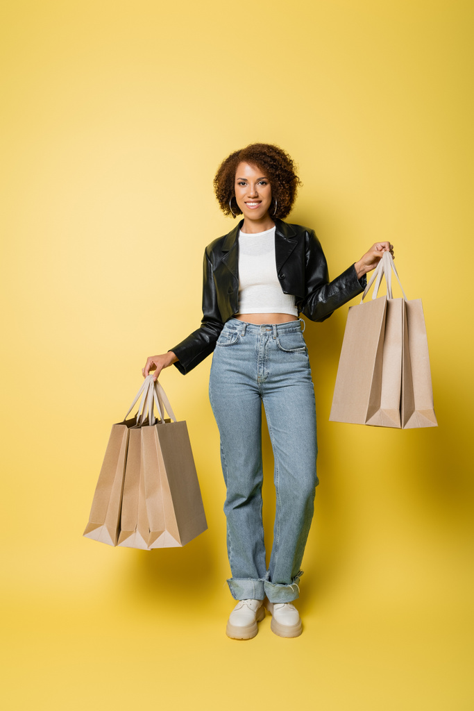 volle Länge positive afrikanisch-amerikanische Frau in stylischer Lederjacke mit Einkaufstaschen mit Einkäufen auf gelb - Foto, Bild