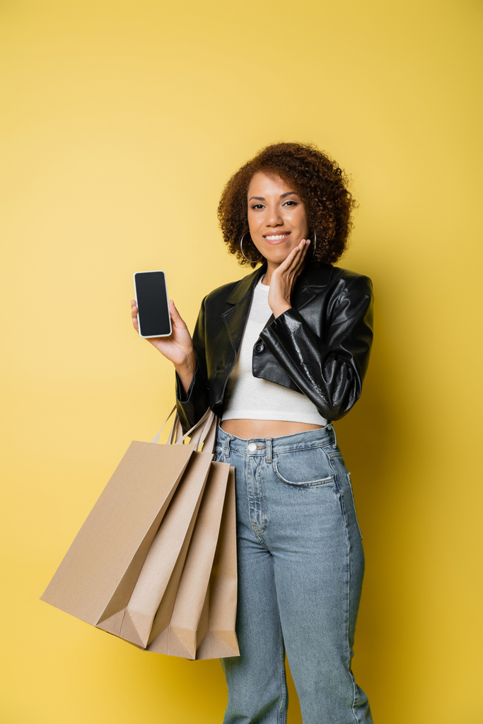 sorrindo mulher afro-americana em jaqueta de couro segurando sacos de compras e telefone celular com tela em branco no amarelo - Foto, Imagem