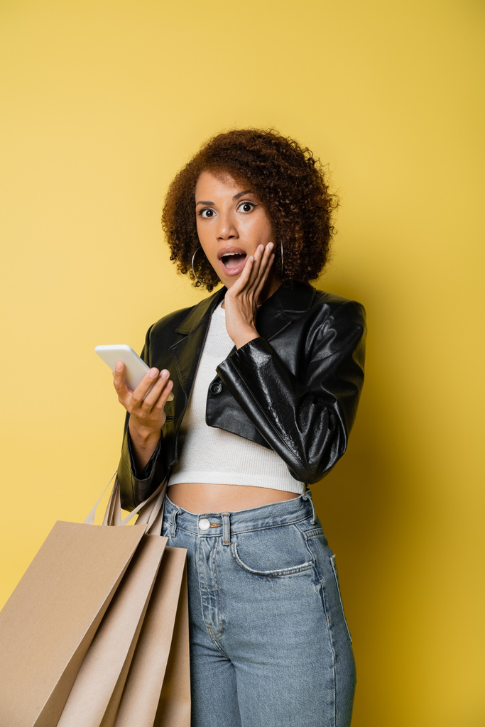 chocado Africano americano mulher em jaqueta de couro segurando sacos de compras e telefone celular no amarelo - Foto, Imagem