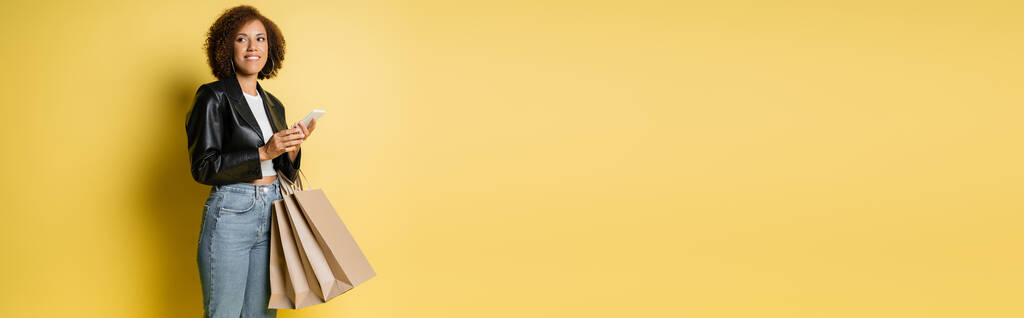 tyytyväinen afrikkalainen amerikkalainen nainen tyylikäs nahkatakki tilalla ostoskassit ja matkapuhelin keltainen, banneri - Valokuva, kuva