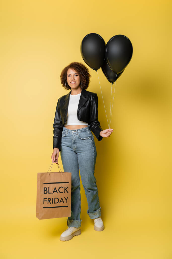 longitud completa de la sonriente mujer afroamericana en chaqueta de cuero con bolsa de compras de viernes negro y globos oscuros en amarillo - Foto, Imagen