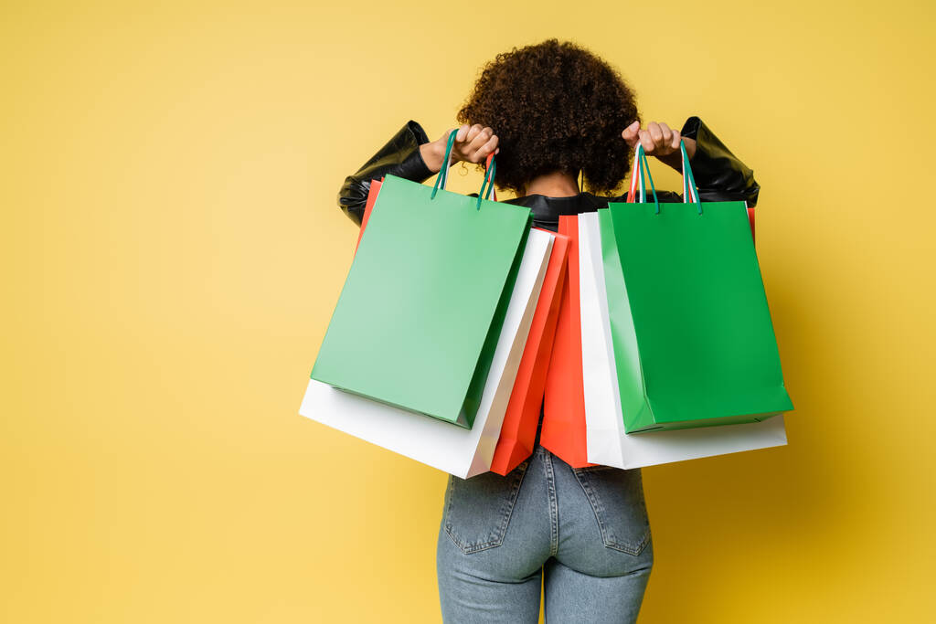 back view of curly african american woman in blue jeans holding colorful black friday shopping bags on yellow - Photo, Image