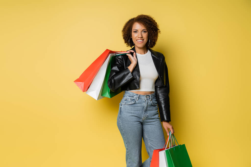 pozitivní africká americká žena v džínách a kožené bundě drží barevné černé pátek nákupní tašky na žluté  - Fotografie, Obrázek