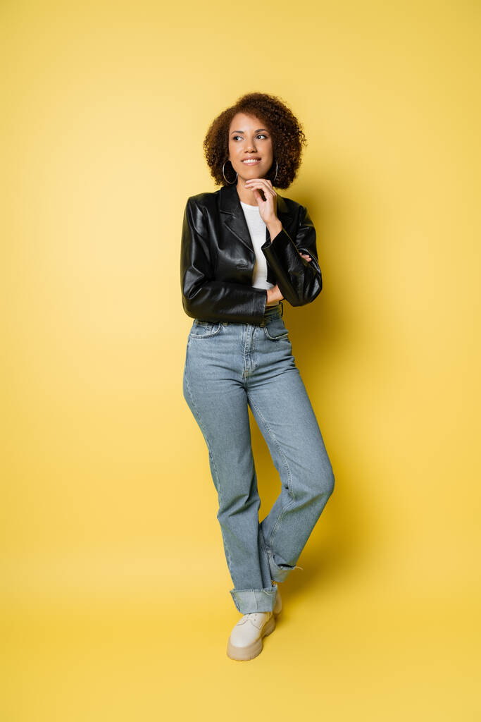 longitud completa de la sonriente mujer afroamericana en jeans y chaqueta de cuero posando en amarillo  - Foto, Imagen