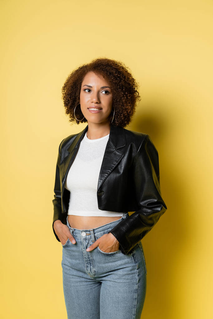smiling african american woman in jeans and leather jacket posing with hands in pockets on yellow  - Photo, Image
