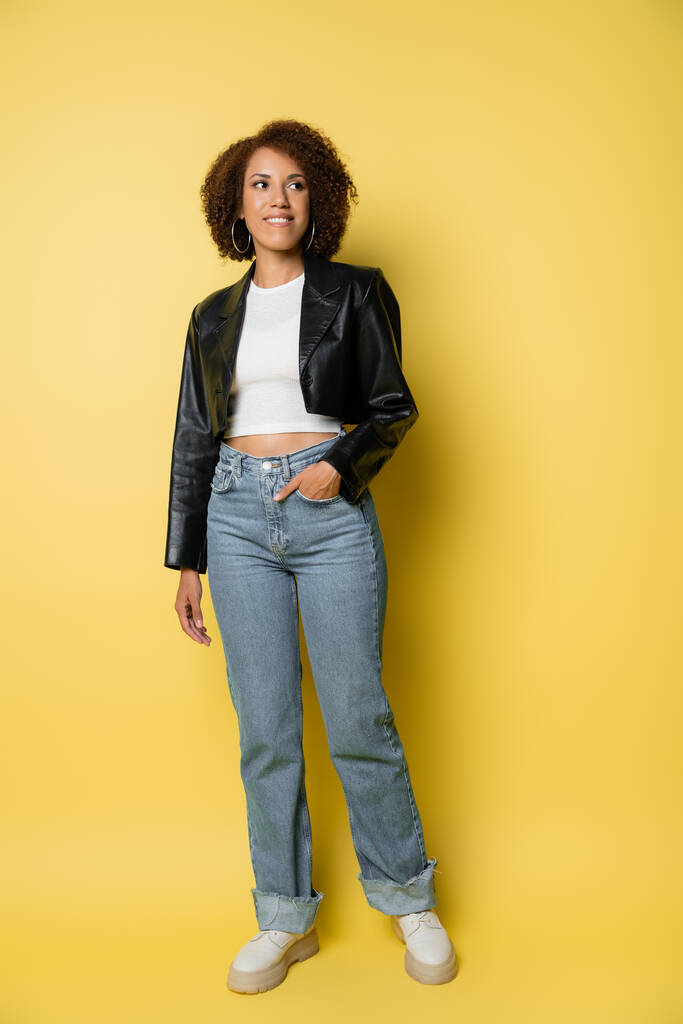 pełna długość pozytywnej afro-amerykańskiej kobiety w dżinsach i skórzanej kurtce pozującej z dłonią w kieszeni na żółtym  - Zdjęcie, obraz