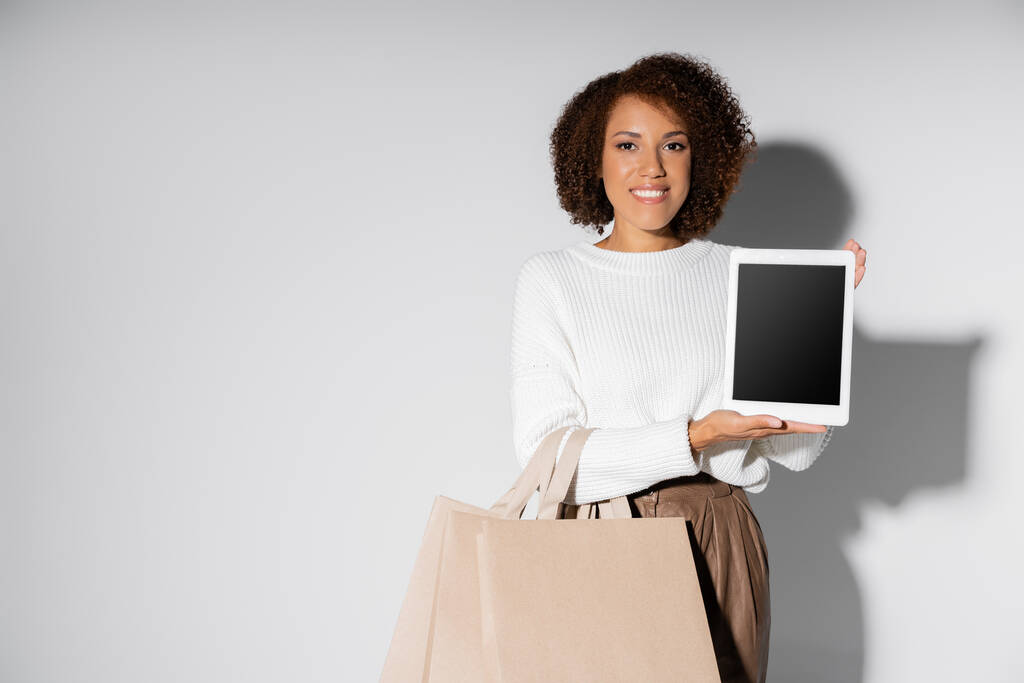 boldog afro-amerikai nő őszi ruhában gazdaság bevásárló táskák és digitális tabletta üres képernyő szürke - Fotó, kép