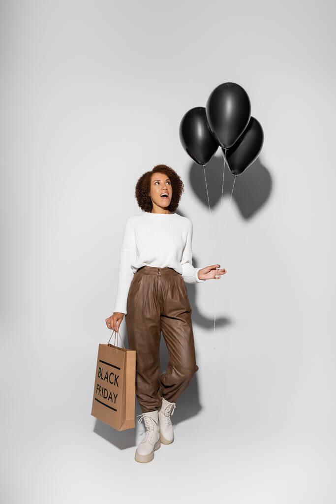teljes hossza meglepett afro-amerikai nő őszi ruhában gazdaság bevásárló táska és fekete lufik szürke - Fotó, kép