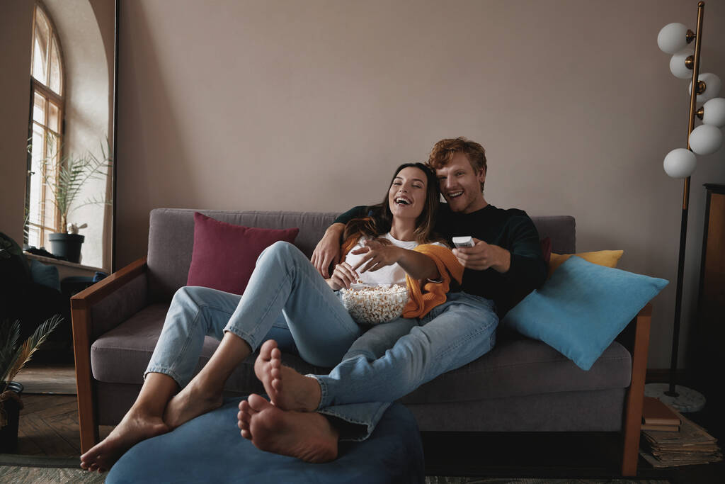 一緒に家でソファでリラックスしながら、美しい若いカップルはテレビを見て笑って - 写真・画像