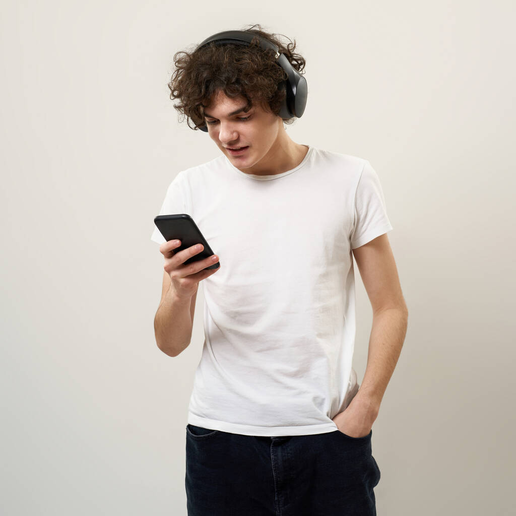 Valkoihoinen teini-ikäinen poika, jolla on käsi taskussa älypuhelimella ja kuuntelemalla musiikkia kuulokkeilla. Zoomer-sukupolven kihara kaveri. Nykyaikainen nuoren elämäntapa. Valkoinen tausta. Studion kuvaukset. Kopioi tila - Valokuva, kuva