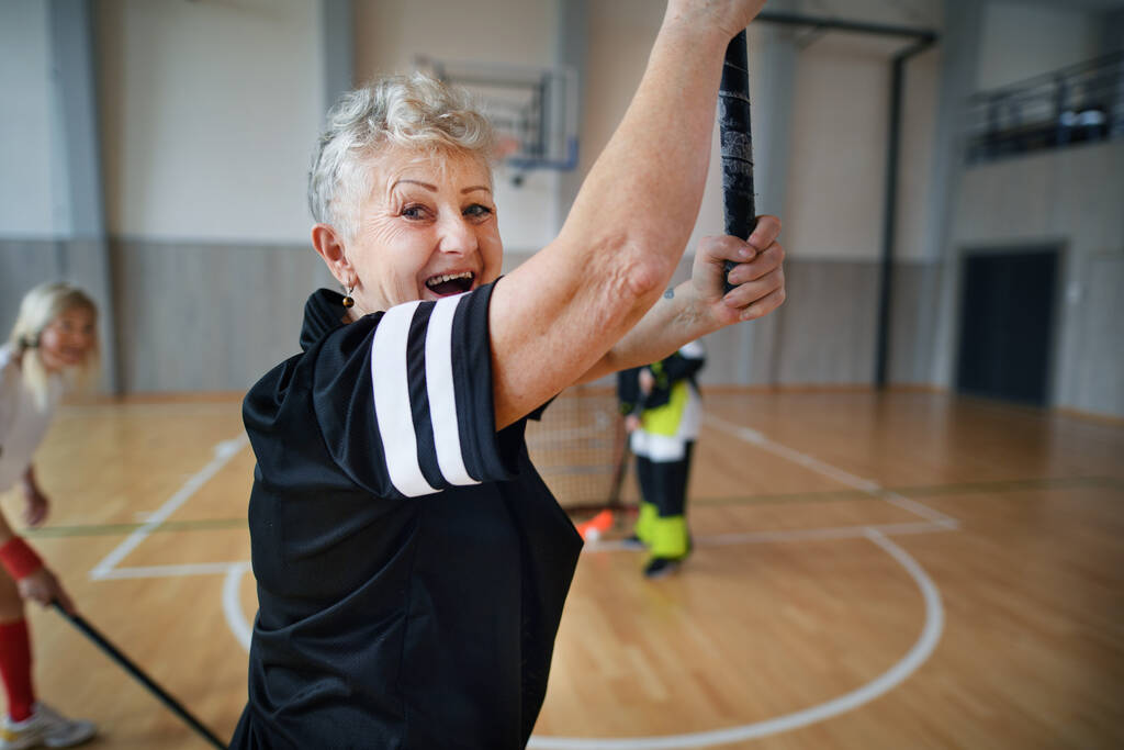 Возбужденная старшая женщина играет в флорбол в спортзале с друзьями. - Фото, изображение
