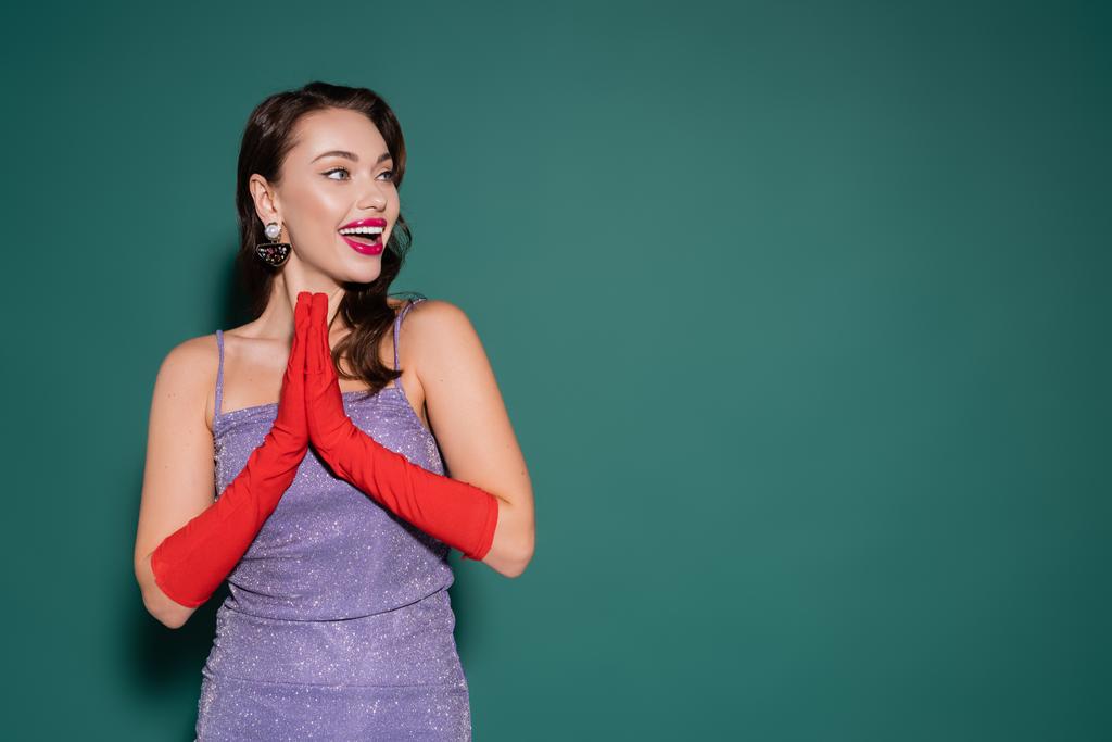 excited young woman in red gloves and purple dress gesturing while looking away on green - Photo, Image