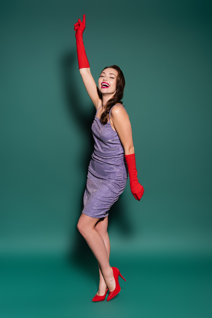 volledige lengte van charmante jonge vrouw in paarse jurk en handschoenen lachen op groen - Foto, afbeelding