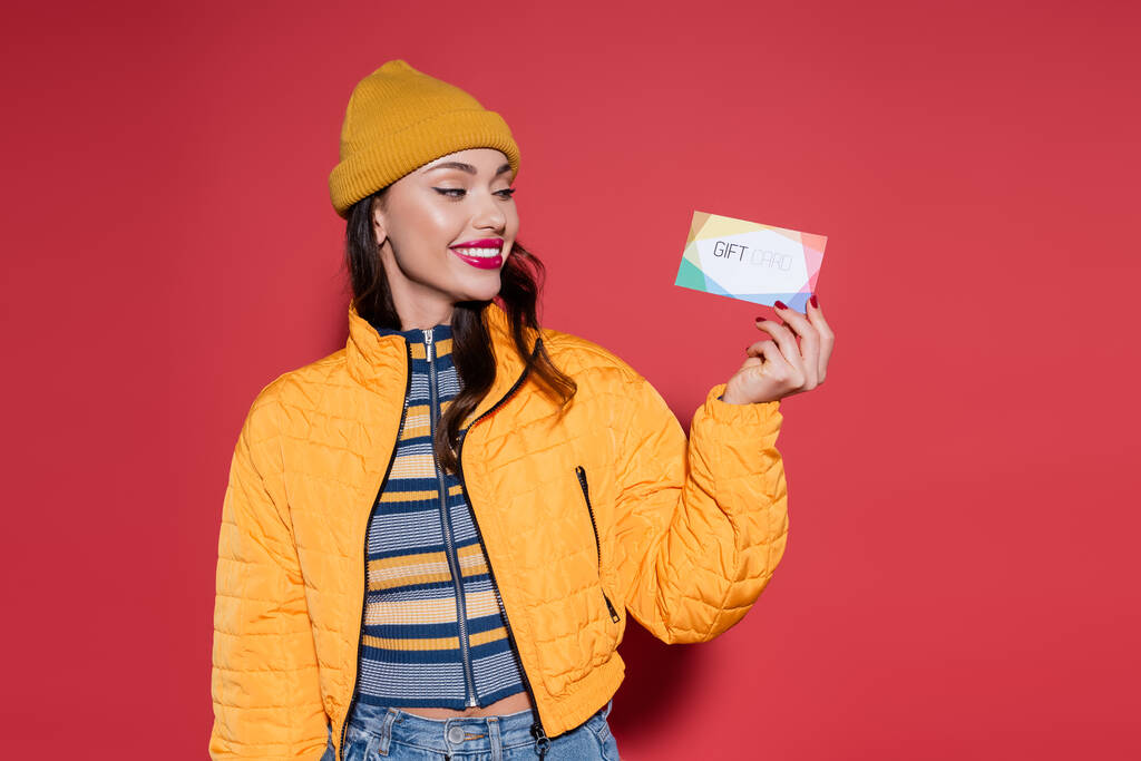 happy young woman in beanie hat and orange puffer jacket holding gift card on red - Photo, Image