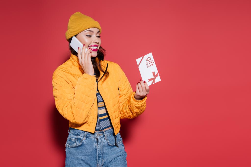 gelukkig vrouw in beanie hoed en oranje kogelvrij vest met gift card terwijl het praten op de smartphone op rood - Foto, afbeelding
