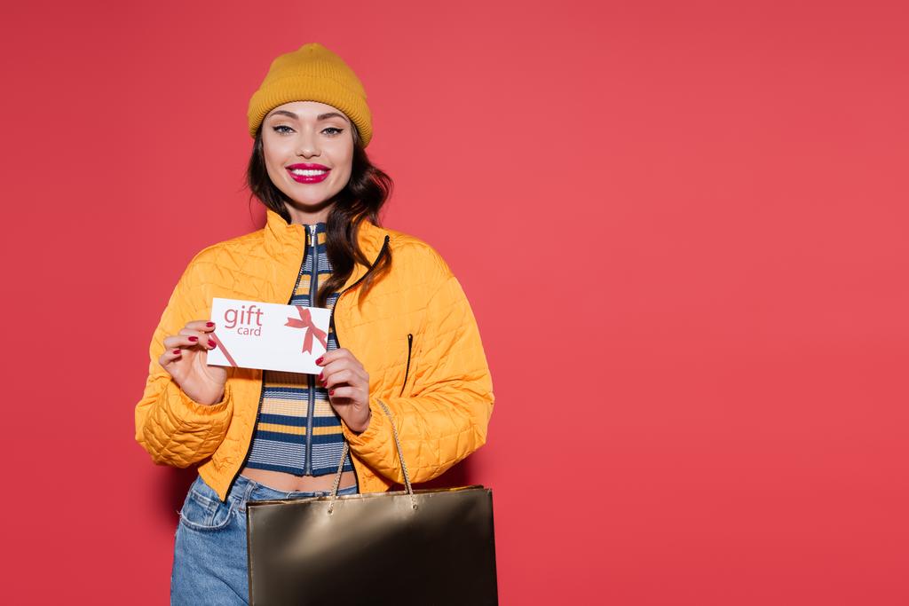 χαρούμενη νεαρή γυναίκα σε πορτοκαλί καπέλο beanie κρατώντας κάρτα δώρο και τσάντα για ψώνια σε κόκκινο - Φωτογραφία, εικόνα