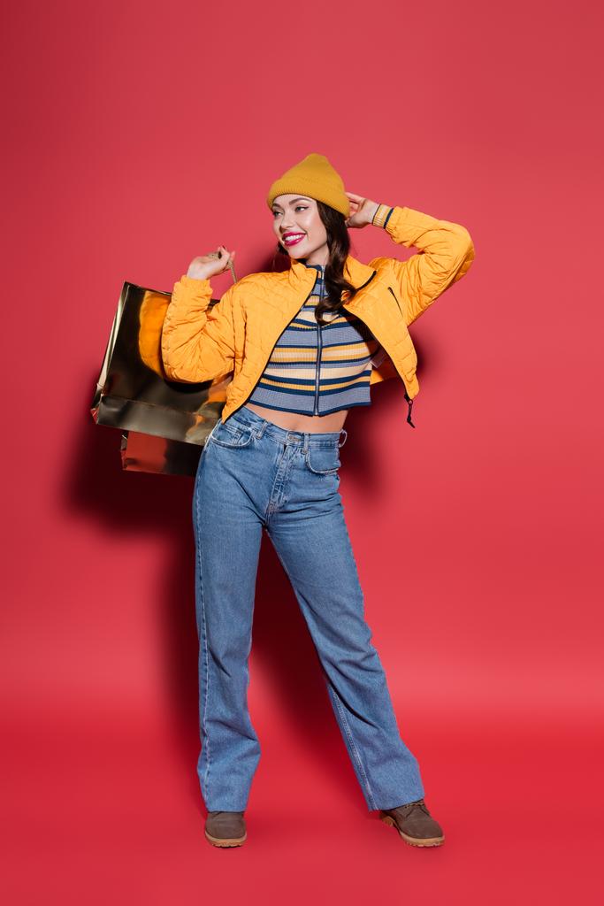 comprimento total da jovem mulher positiva em chapéu de gorro laranja e casaco de puffer segurando saco de compras no vermelho - Foto, Imagem