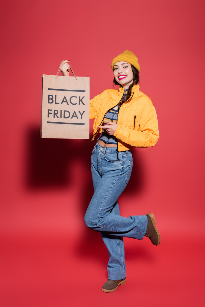 in voller Länge glückliche junge Frau mit Mütze und Pufferjacke zeigt auf Einkaufstasche mit schwarzem Freitag-Schriftzug auf rot - Foto, Bild