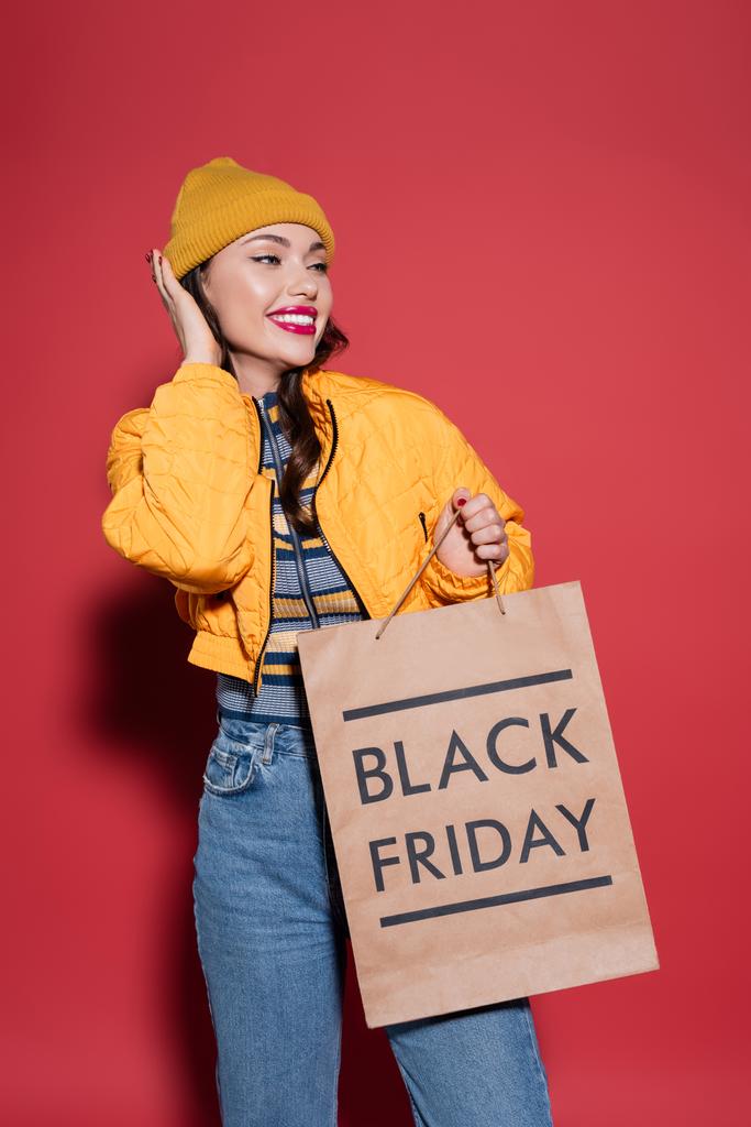gelukkig jong vrouw in oranje muts en kogelvrije vest holding shopping bag met zwarte vrijdag belettering op rood - Foto, afbeelding