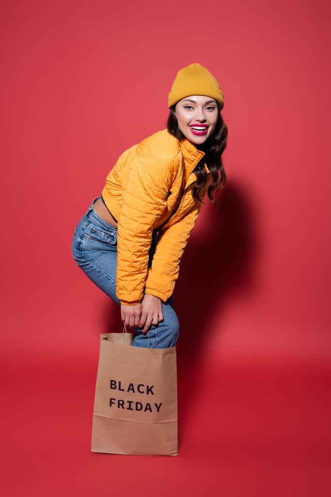 in voller Länge fröhliche junge Frau mit Mütze und Pufferjacke, die in einer Einkaufstasche mit schwarzem Freitag-Schriftzug auf Rot steht - Foto, Bild