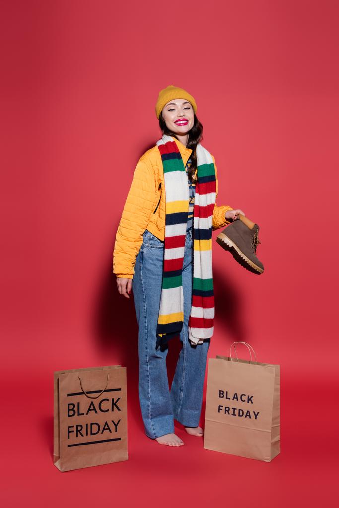 mujer descalza en chaqueta hinchable de pie con bota cerca de bolsas de compras con letras de viernes negro en rojo - Foto, imagen