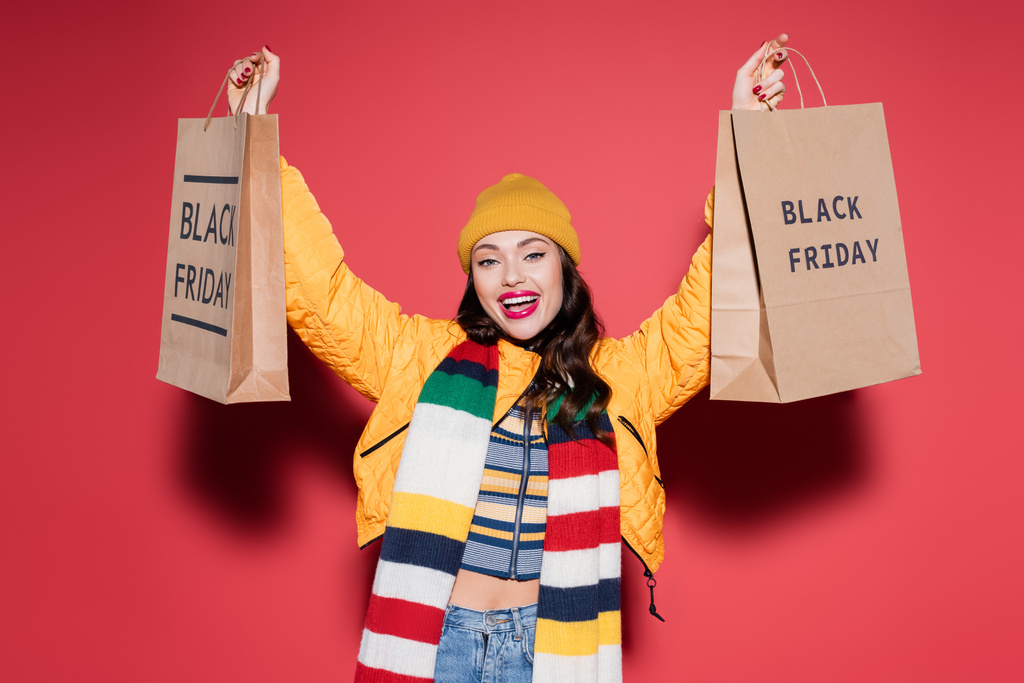 verbaasd vrouw in muts en sjaal houden boodschappentassen met zwarte vrijdag belettering op rood  - Foto, afbeelding
