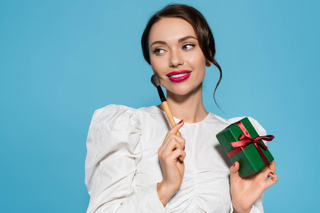 Beyaz bluzlu neşeli genç bir kadın hediye paketi ve kozmetik fırça taşıyor. Mavi renkte izole edilmiş. - Fotoğraf, Görsel