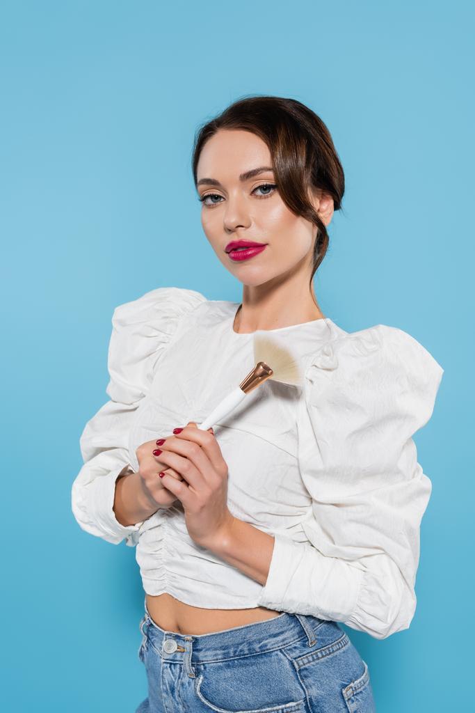 pretty young woman in white blouse holding cosmetic brush isolated on blue - Photo, Image