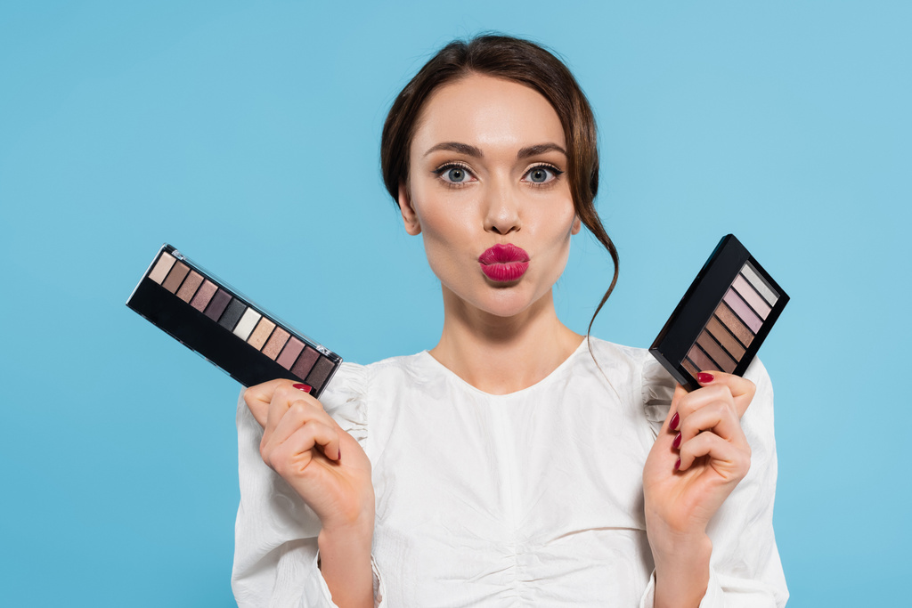 pretty young woman holding eye shadow palettes and pouting lips isolated on blue  - Photo, Image