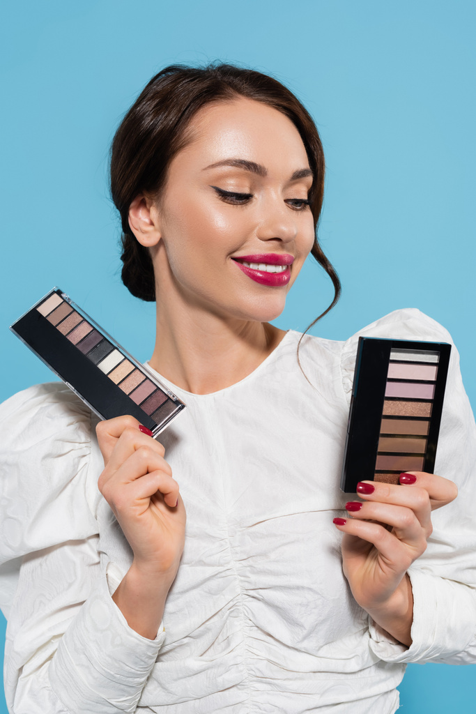 cheerful young woman holding eye shadow palettes isolated on blue  - Photo, Image