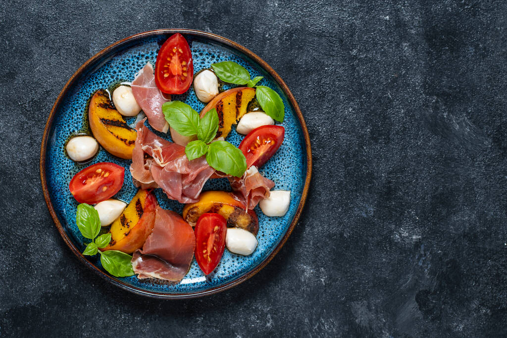Grilled peach salad with mozzarella, prosciutto ham, red tomato, green basil and soy sauce. Fresh gourmet salad in plate on black background, close up, top view, copy space - Photo, Image