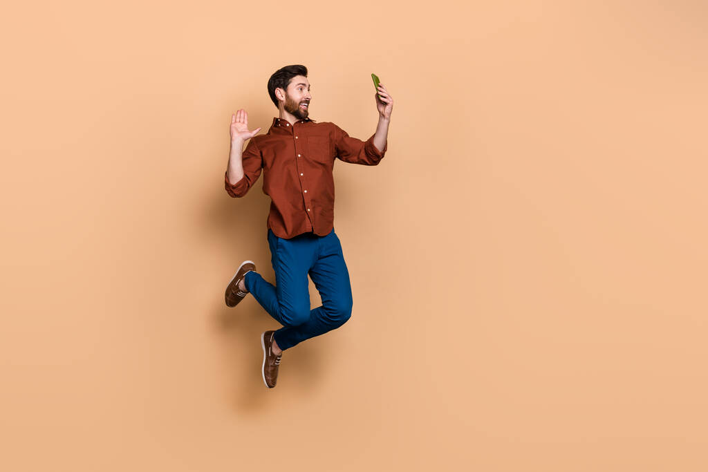 Volledige lengte portret van knappe man springen arm palm zwaaien hi communiceren video chat geïsoleerd op beige kleur achtergrond. - Foto, afbeelding