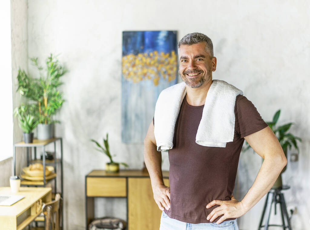 笑顔の幸せな運動中の高齢者の白髪の男は、家庭内で肩にタオルで立っている。自宅でハンサムな成熟した男。人の健康、朝の手順。シャワーマンの後.  - 写真・画像