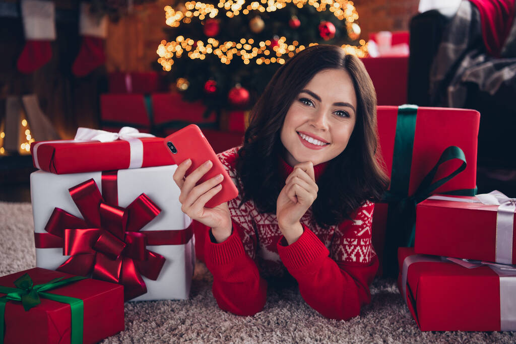 Foto von schönen fröhlichen Mädchen Hand berühren Kinn halten Telefon Verlegung Teppich Boden Geschenkbox drinnen. - Foto, Bild