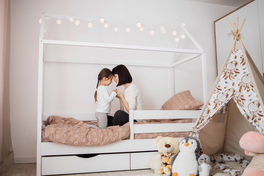 Ein nettes Mädchen und ihre Mutter umarmen sich und genießen den Sonntagmorgen. Gute Zeit zu Hause. Kind wacht aus dem Schlaf auf Familie spielt auf dem Bett im Schlafzimmer. - Foto, Bild