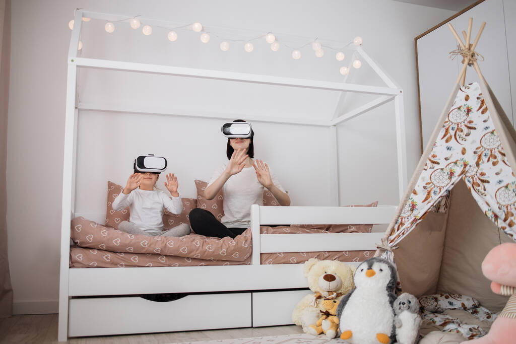 Attractive smiling caucasian woman with her cute little daughter, using virtual reality devices and watching excited movie or playing video game sitting on bed. - Photo, Image