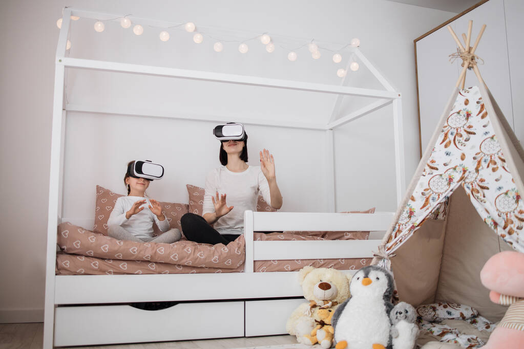 Neue digitale Technologie. Freudig schöne kaukasische Mutter und Tochter lächeln, während sie Virtual-Reality-Brille zu Hause auf skandinavischem Holzbett sitzen. - Foto, Bild