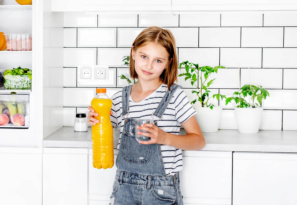 Preteen girl with orange juice bottle and glass at kitchen. Pretty child kid with vitamin fruit drink at home - Fotó, kép