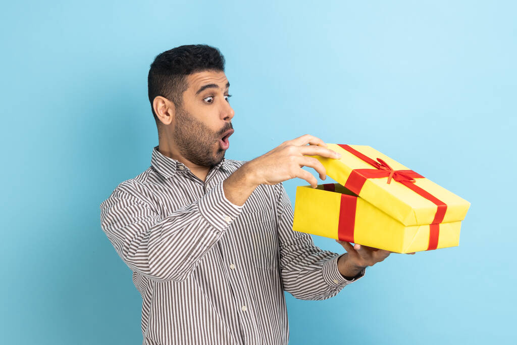Portrait d'homme d'affaires curieux surpris regardant dans une boîte-cadeau, ouvrant le cadeau et regardant à l'intérieur avec intérêt, portant une chemise rayée. Studio intérieur tourné isolé sur fond bleu. - Photo, image