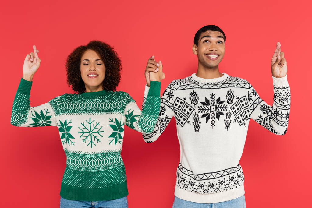 Αφροαμερικανό ζευγάρι σε πουλόβερ με χειμερινό σχέδιο κρατώντας τα χέρια και σταυρώνοντας τα δάχτυλα απομονώνονται σε κόκκινο - Φωτογραφία, εικόνα