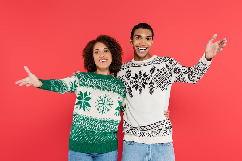 glückliches afrikanisch-amerikanisches Paar in warmen Pullovern mit Winterornament, die Grußgeste isoliert auf rot - Foto, Bild