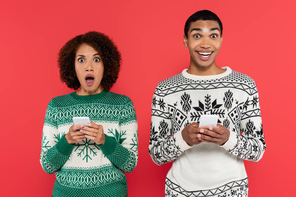 asombrada pareja afroamericana en suéteres cálidos mirando a la cámara mientras sostiene los teléfonos inteligentes aislados en rojo - Foto, Imagen