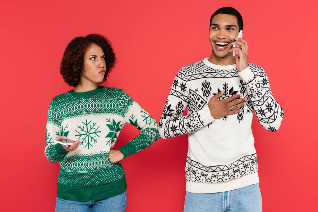 beleidigte Afroamerikanerin steht mit Hand auf Hüfte neben lachendem Freund und spricht auf Smartphone isoliert auf Rot - Foto, Bild