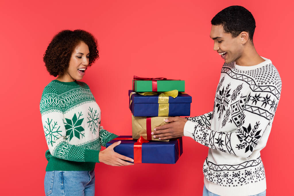asombrada pareja afroamericana en suéteres cálidos con adorno de invierno celebración de regalos de año nuevo aislados en rojo - Foto, Imagen